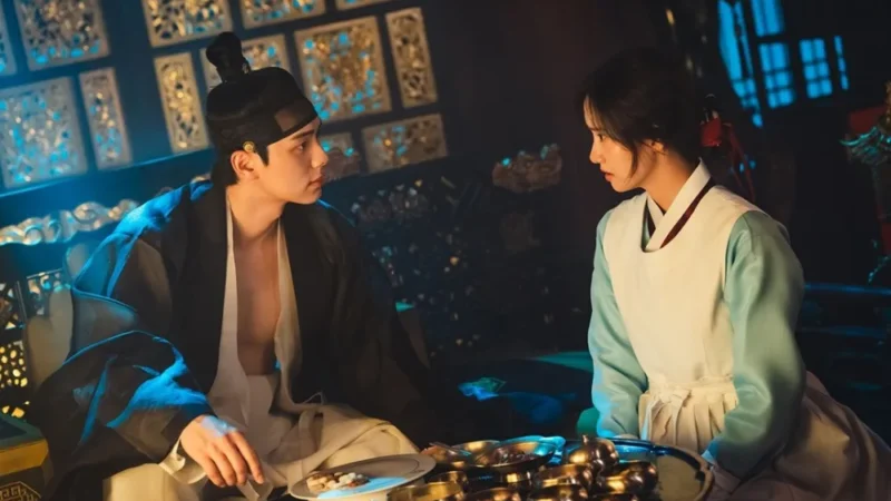 A chef Yeon Ji-yeong e o rei Lee Heon frente a frente diante de uma farta mesa de pratos no palácio de Joseon, em luz cinematográfica azul e dourada